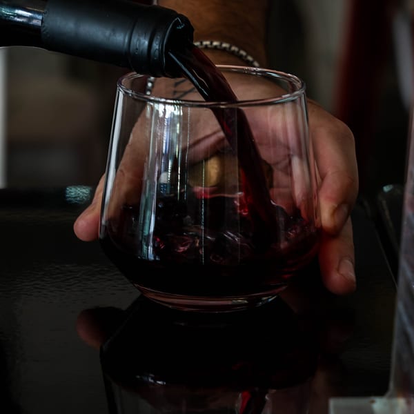 Hand pouring red wine from a bottle into a stemless glass on a dark surface.