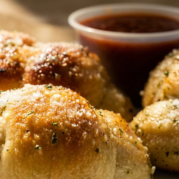 Golden-brown garlic knots dusted with parmesan cheese and herbs, with a side of marinara sauce.