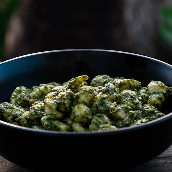 A black bowl filled with gnocchi coated in green pesto sauce, on a rustic wooden surface.