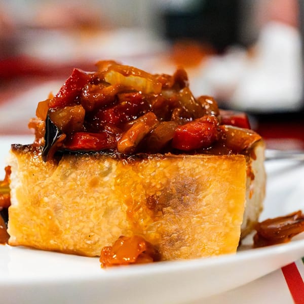 Close-up of a bruschetta topped with a savory red and orange vegetable mixture on a white plate.